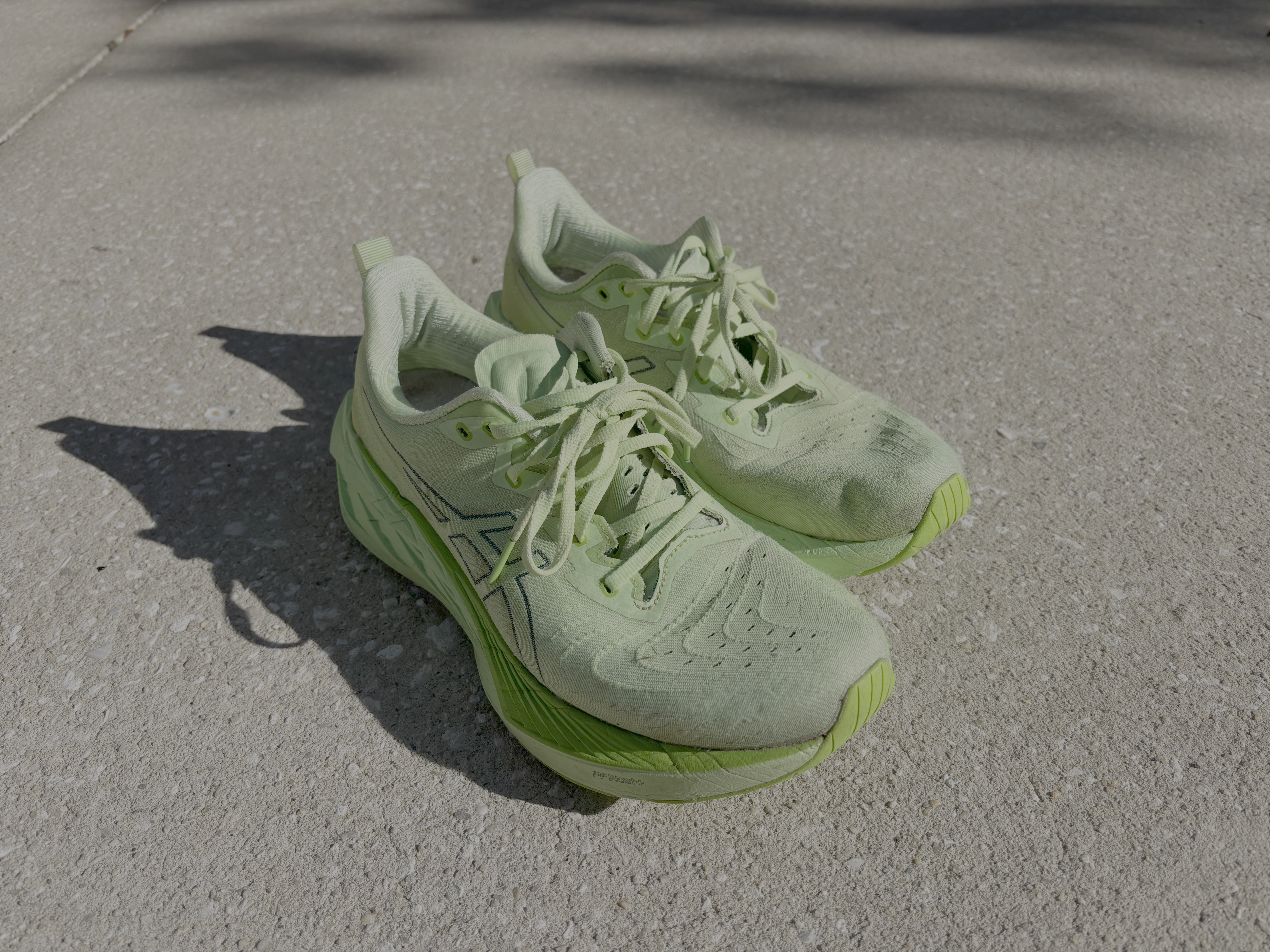 A pair of well-worn ASICS NovaBlast running shoes in bright green, placed on a concrete sidewalk. The shoes show visible signs of use, with dirt and scuff marks on the mesh upper and outsole. Shadows from the shoes are cast onto the pavement under bright sunlight.